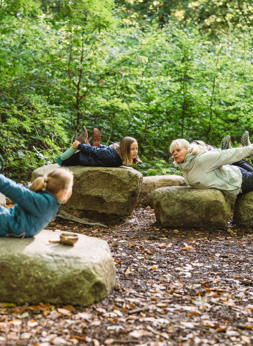 Kinder und ein Erwachsener liegen auf gro&szlig;en Steinen im Wald und machen gemeinsam Yoga-&Uuml;bungen, umgeben von dichten B&auml;umen und Bl&auml;ttern.