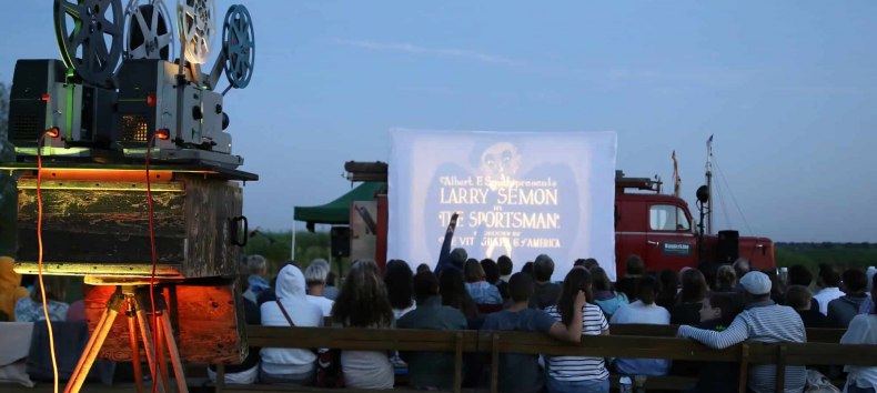 Open-Air-Kinoabend des Wanderkinos Althagen in Ahrenshoop mit einem historischen 16mm- oder 35mm-Filmprojektor im Vordergrund und Publikum vor der Leinwand. // © Tobias Rank Open-Air-Kinoabend des Wanderkinos Althagen in Ahrenshoop mit einem historischen 16mm- oder 35mm-Filmprojektor im Vordergrund und Publikum vor der Leinwand. // © Tobias Rank