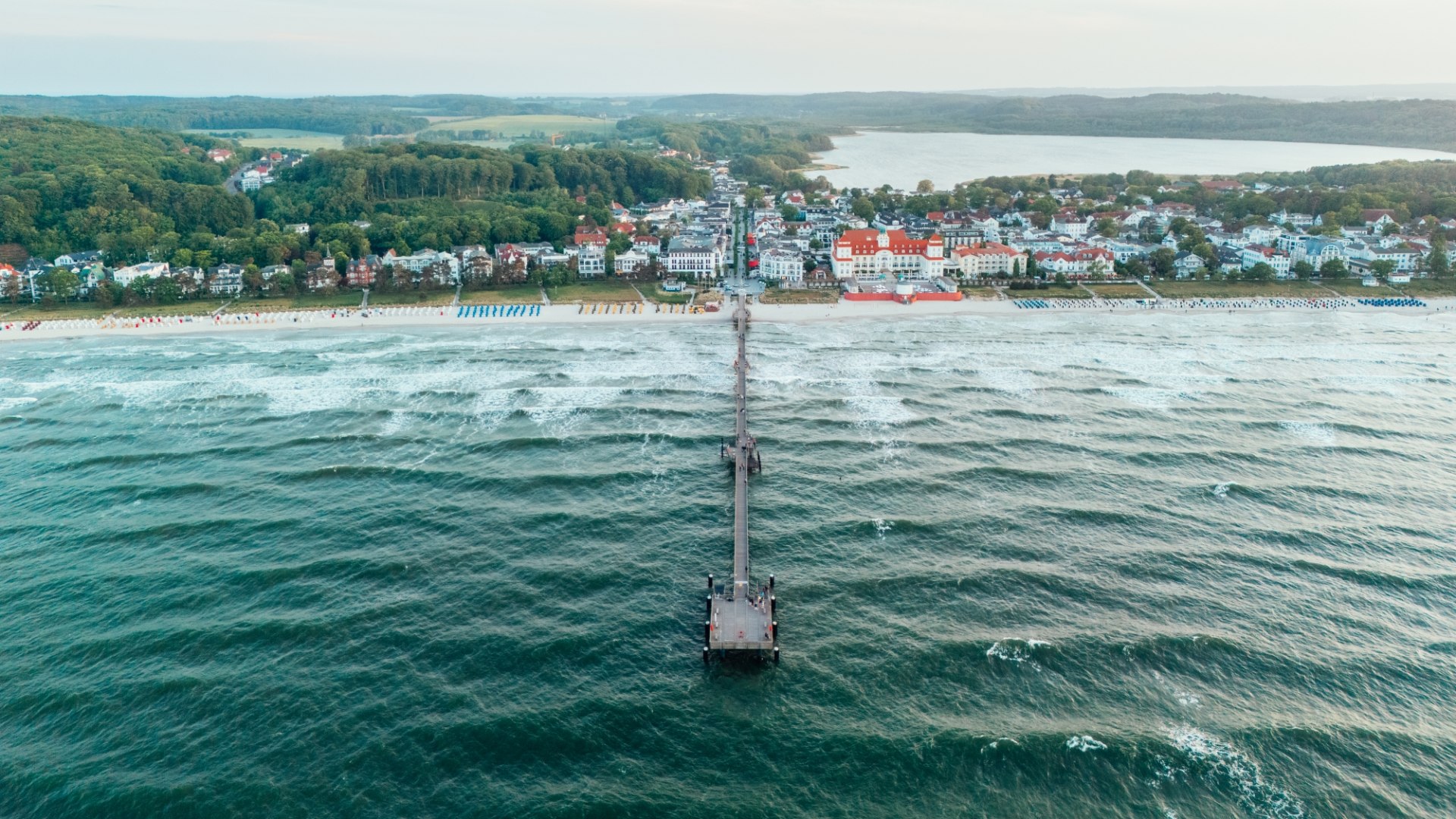 Luftaufnahme der Seebrücke in Binz auf Rügen, die weit in die Ostsee hineinragt. Dahinter liegt Binz mit seiner prächtigen Bäderarchitektur, eingebettet in eine hügelige Landschaft.