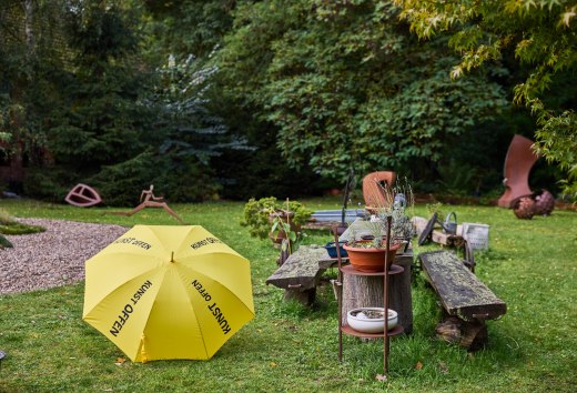 Skulpturengarten mit gelbem KunstOffen-Schirm und Metallskulpturen im Kunst-Werk Kummer. Davor liegt ein gelber Regenschirm mit der Aufschrift "KunstOffen"