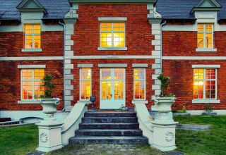 Illuminierter Eingang des Herrenhauses Brookhusen: Historistische Backsteinfassade und einladend leuchtende Fenster in der Abenddämmerung. // © Herrenhaus Brockhusen Illuminierter Eingang des Herrenhauses Brookhusen: Historistische Backsteinfassade und einladend leuchtende Fenster in der Abenddämmerung. // © Herrenhaus Brockhusen