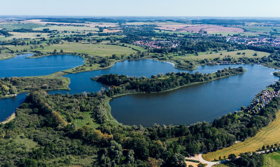 Luftaufnahme vom Teterower See mit Burgwallinsel, gr&uuml;nen Ufern, Feldern und Teterow im Hintergrund.