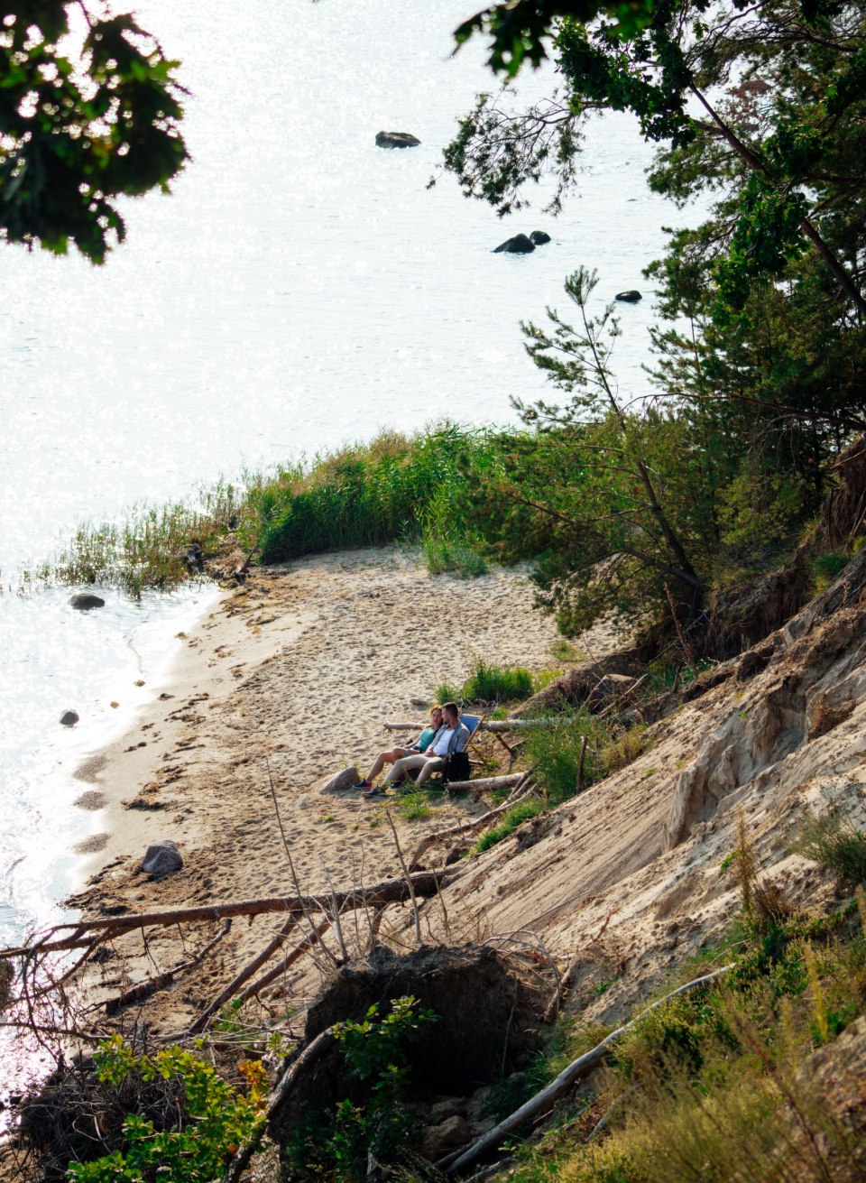Ein kleiner Sandstrand im Biosphärenreservat Südost-Rügen, umgeben von steilen Uferhängen und dichter Vegetation. Zwei Personen sitzen entspannt auf einer Bank, mit Blick auf das glitzernde Wasser, das von vereinzelten Felsen durchzogen ist. Eine ruhige und abgeschiedene Atmosphäre lädt zum Verweilen ein. // Ein idyllischer Rückzugsort am Strand des Biosphärenreservats Südost-Rügen – Ruhe und Natur pur. // © MV-T/Petermann Ein kleiner Sandstrand im Biosphärenreservat Südost-Rügen, umgeben von steilen Uferhängen und dichter Vegetation. Zwei Personen sitzen entspannt auf einer Bank, mit Blick auf das glitzernde Wasser, das von vereinzelten Felsen durchzogen ist. Eine ruhige und abgeschiedene Atmosphäre lädt zum Verweilen ein.