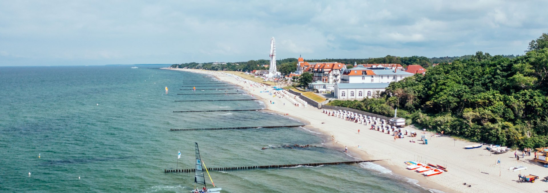 Luftaufnahme vom Strand in K&uuml;hlungsborn mit Buhnen, Strandk&ouml;rben, Riesenrad und Segelboot auf der Ostsee.
