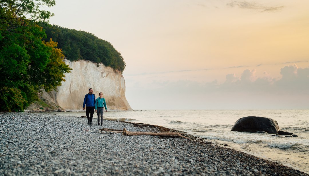 Een koppel wandelt langs de voet van de beroemde krijtrotsen op het eiland R&uuml;gen, terwijl de zonsondergang de hemel in zachte kleuren oplicht.