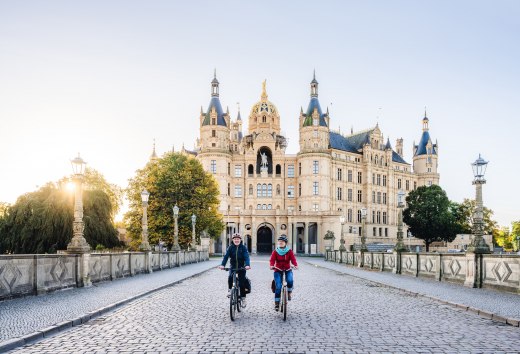 Zwei Radfahrer &uuml;berqueren eine Br&uuml;cke vor dem Schweriner Schloss, beleuchtet von der goldenen Morgensonne.