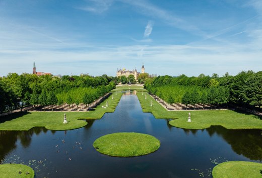 Baroktuin van Schwerin met symmetrische gracht, waterpartijen en kasteel op de achtergrond onder een blauwe hemel. // De paleistuinen van Schwerin nodigen je uit om diep adem te halen - barokke waterpartijen weerspiegelen het sprookjesachtige paleis, terwijl lanen met lindebomen voor schaduw zorgen. Ontdek waar architectuur en natuur harmonieus samensmelten. // © MV-T/Gänsicke Baroktuin van Schwerin met symmetrische gracht, waterpartijen en kasteel op de achtergrond onder een blauwe hemel.