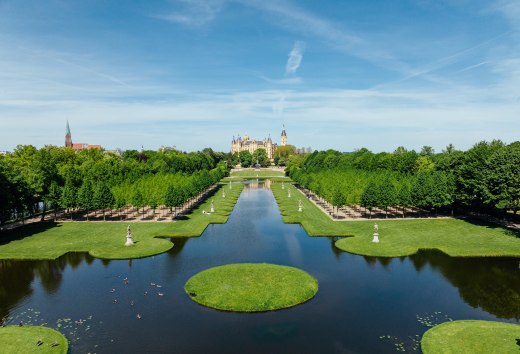 Barockgarten Schwerin mit symmetrischem Kanal, Wasserflächen und Schloss im Hintergrund unter blauem Himmel. // Der Schweriner Schlossgarten lädt zum Durchatmen ein – barocke Wasserspiele spiegeln das märchenhafte Schloss, während Lindenbaumalleen Schatten spenden. Entdecken, wo Architektur und Natur harmonisch verschmelzen. // © MV-T/Gänsicke Barockgarten Schwerin mit symmetrischem Kanal, Wasserflächen und Schloss im Hintergrund unter blauem Himmel.