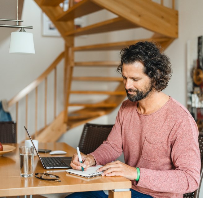 Ein Mann sitzt an einem Holztisch in einem Ferienhaus, arbeitet am Laptop und macht Notizen &ndash; ideale Bedingungen f&uuml;r eine Workation auf der Insel Poel.