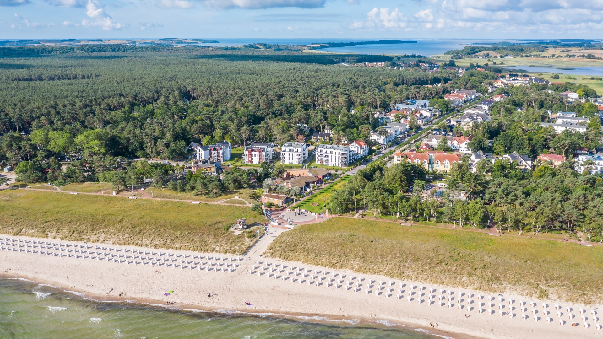Luftaufnahme des Ostseebads Baabe auf der Insel R&uuml;gen: Strand mit Strandk&ouml;rben, D&uuml;nen, moderne Villen und Ferienh&auml;user, umgeben von dichten W&auml;ldern und Blick auf die Boddenlandschaft.