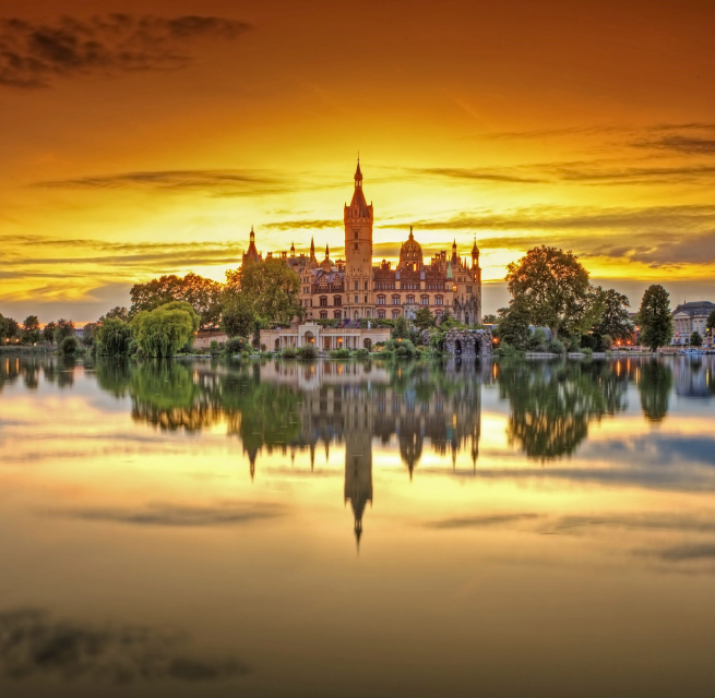 Das Schweriner Schloss wird von den letzten Sonnenstrahlen umrahmt. Der Sonnenuntergang färbt den Himmel orange. Im ruhigen See spiegelt sich das Schweriner Schloss.