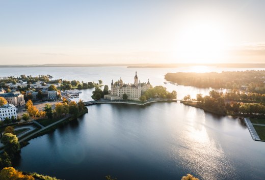Die Residenz auf der Schlossinsel thront zwischen Wasser und Himmel. Morgenlicht streift die Türme des Schweriner Schlosses, während sich die Silhouette im stillen See spiegelt. Geschichte trifft auf Weite – ein Ort, der Raum zum Durchatmen lässt und gleichzeitig die Blicke festhält., © MV-T/Gross Schweriner Schloss auf Insel im See bei Sonnenaufgang, umgeben von Bäumen und Wasserflächen.