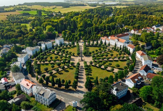 Luftaufnahme des Circus in Putbus auf Rügen, einem kreisförmigen Platz mit symmetrisch angeordneten Bäumen und klassizistischen Gebäuden, umgeben von grüner Natur und ländlicher Landschaft. // Der historische Circus in Putbus auf der Insel Rügen – ein beeindruckendes klassizistisches Bauensemble inmitten einer gepflegten Parklandschaft, ideal für Spaziergänge und kulturelle Entdeckungen. // © MV-T/Petermann Luftaufnahme des Circus in Putbus auf Rügen, einem kreisförmigen Platz mit symmetrisch angeordneten Bäumen und klassizistischen Gebäuden, umgeben von grüner Natur und ländlicher Landschaft.