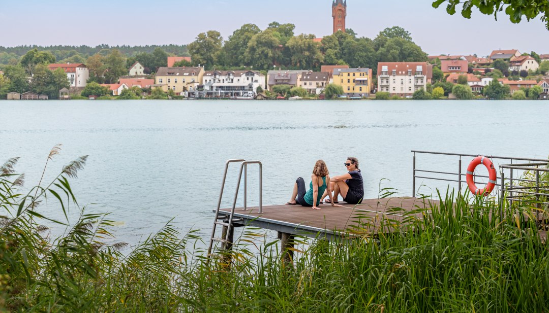 Zwei Frauen sitzen auf einem Steg und blicken dabei auf einen See