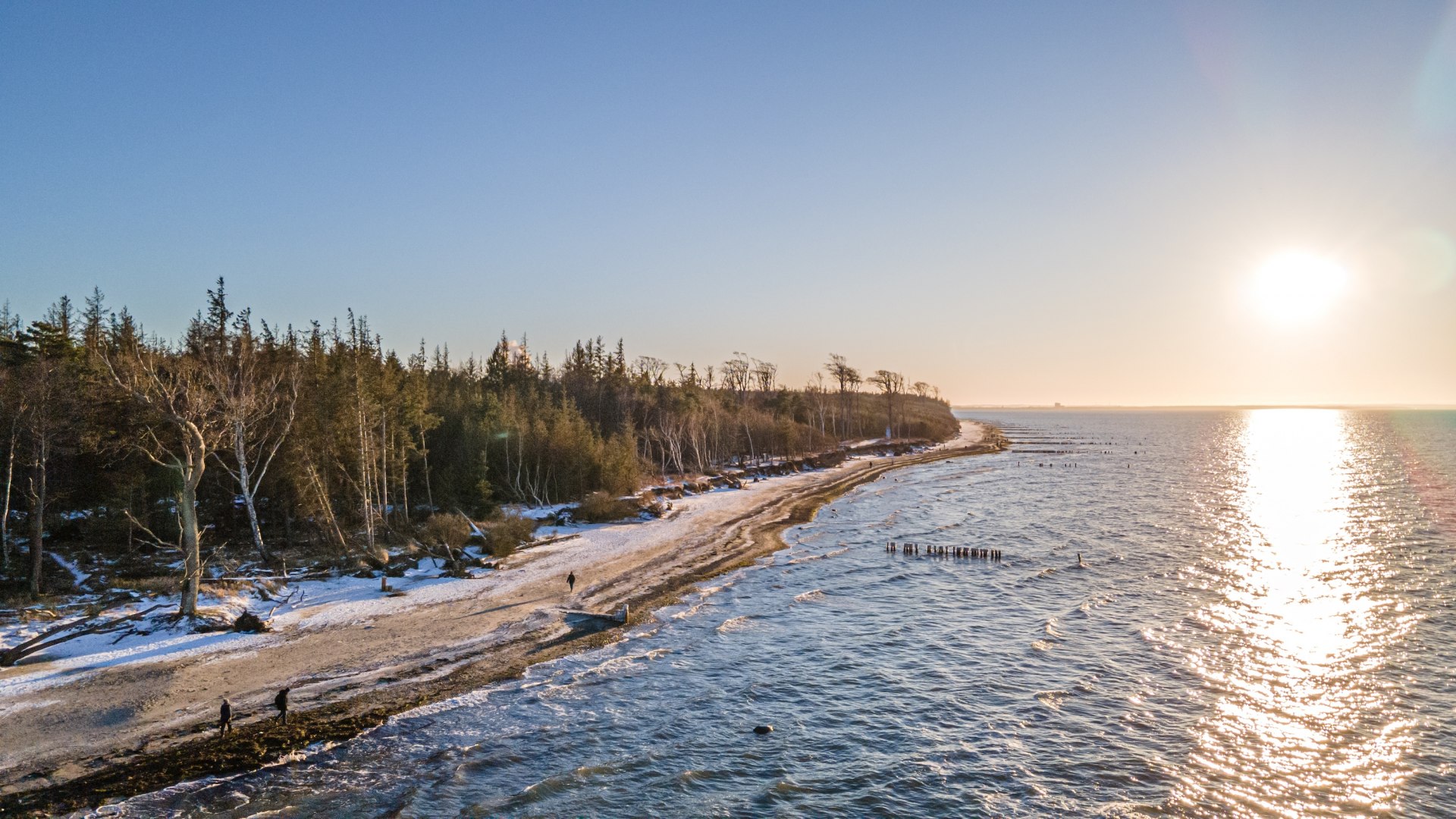 Winterlicher Strand bei Torfbr&uuml;cke Graal-M&uuml;ritz mit schneebedeckten B&auml;umen, glitzernder Ostsee und einem Sonnenuntergang am Horizont.