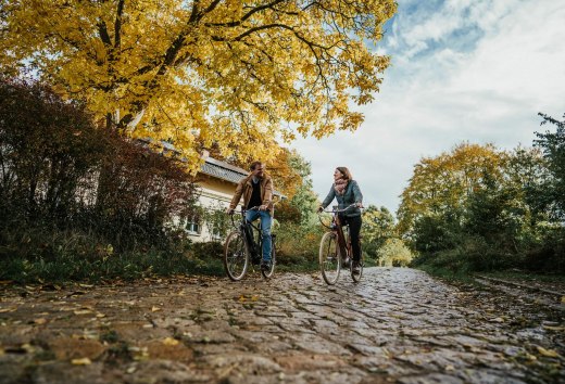 Unterwegs mit dem Fahrrad am Gut Pohnstorf im Herbst, © 1000seen Zwei Personen fahren mit dem Fahrrad im Herbst auf Kopfsteinpflaster beim Gut Pohnstorf.