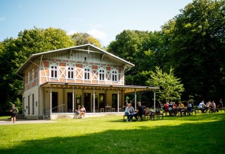 Ein helles Gebäude mit einer Terrasse, vor dem mehrere Personen auf Bänken auf einer grünen Wiese sitzen. // © Nationalpark-Zentrum KÖNIGSSTUHL / Dan Petermann Ein helles Gebäude mit einer Terrasse, vor dem mehrere Personen auf Bänken auf einer grünen Wiese sitzen. // © Nationalpark-Zentrum KÖNIGSSTUHL / Dan Petermann