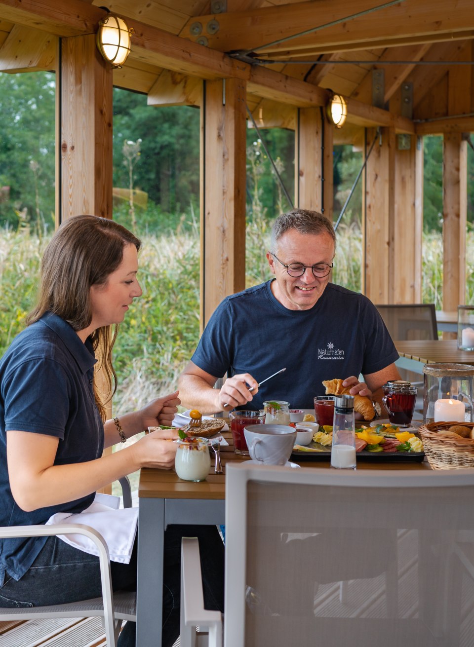EIn paar sitzt an einem reichhaltig gedeckten Frühstückstisch im Naturhafen Krummin. // Erst mal "regional" frühstücken im neuen Glashaus: Frank Schmidt und Fenja Saathoff vom Naturhafen Krummin // © MV-T/Tiemann EIn paar sitzt an einem reichhaltig gedeckten Frühstückstisch im Naturhafen Krummin.