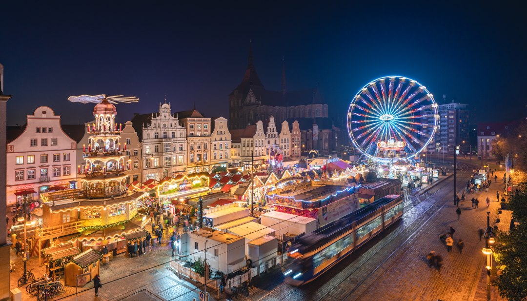 Blick auf den festlich beleuchteten Weihnachtsmarkt in Rostock mit Riesenrad und historischen Geb&auml;uden bei Nacht.