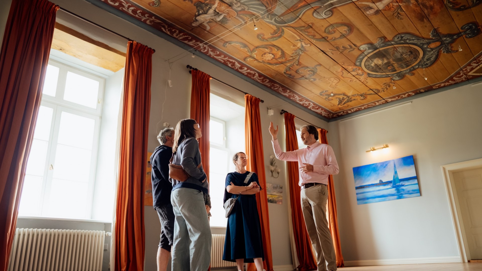 Jannis Achtenhagen erklärt Gästen im Barocksaal des Gutshauses Ludorf die bemalte Decke, hohe Fenster mit Vorhängen. // annis Achtenhagen hebt die Hand, lenkt den Blick zur bemalten Decke und erzählt die nächsten Details – Führung im Barocksaal des Gutshauses Ludorf // © MV-T/Petermann Jannis Achtenhagen erklärt Gästen im Barocksaal des Gutshauses Ludorf die bemalte Decke, hohe Fenster mit Vorhängen.