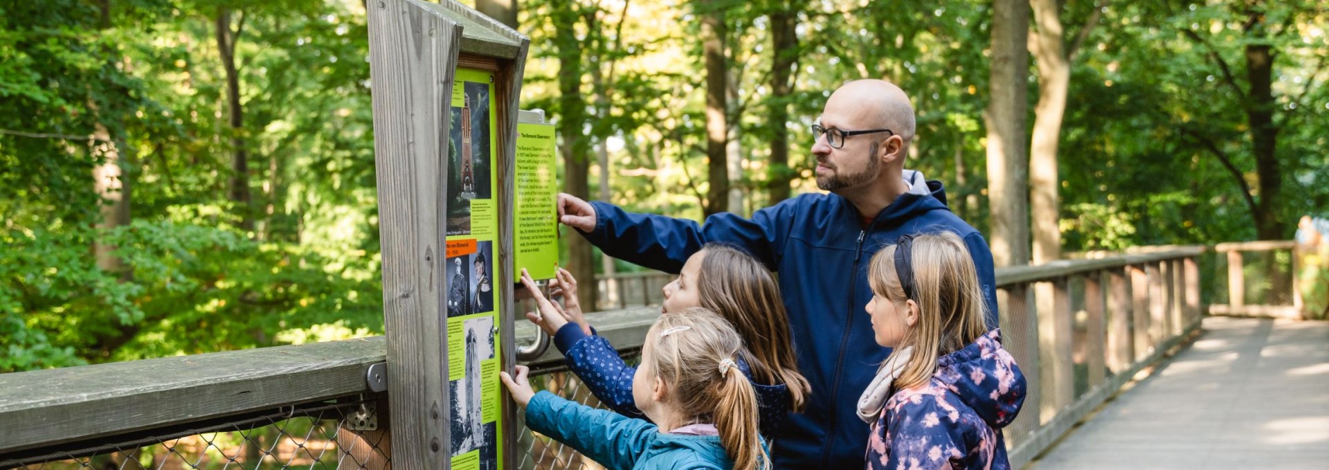 Eine Gruppe von Kindern und ein Erwachsener betrachten eine Infotafel auf einem h&ouml;lzernen Baumwipfelpfad, umgeben von gr&uuml;nen B&auml;umen.