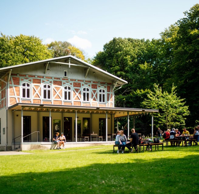 Ein helles Gebäude mit einer Terrasse, vor dem mehrere Personen auf Bänken auf einer grünen Wiese sitzen. // © Nationalpark-Zentrum KÖNIGSSTUHL / Dan Petermann Ein helles Gebäude mit einer Terrasse, vor dem mehrere Personen auf Bänken auf einer grünen Wiese sitzen. // © Nationalpark-Zentrum KÖNIGSSTUHL / Dan Petermann