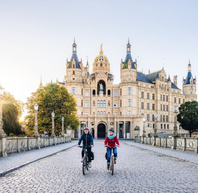 Märchenhafte Morgensonne – Das Schweriner Schloss in herbstlicher Kulisse lädt für die Fahrradtour ein., © TMV/Gross Zwei Radfahrer überqueren eine Brücke vor dem Schweriner Schloss, beleuchtet von der goldenen Morgensonne.