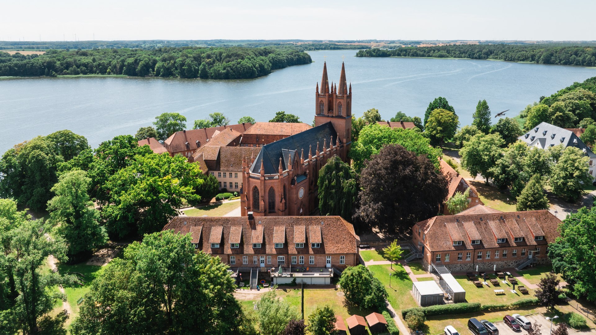 Luftaufnahme vom Kloster Dobbertin mit Backsteinkirche und T&uuml;rmen am See, umgeben von gr&uuml;nen B&auml;umen und Uferwegen.