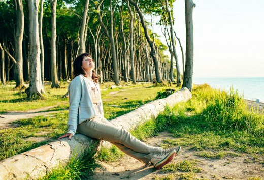 Waldbaden im Gespensterwald - eine Frau sitzt in der Sonne auf einem umgefallenen Baumstamm mit Blick auf die Ostsee // Für positive Effekte auf Körper und Geist sorgt die besondere Beschaffenheit der Küstenregion beim Ostseebad Nienhagen. Denn auf engem Raum hat man ganz verschiedene klimatische Bedingungen – die kühlere Seeseite, den windgeschützten Küstenwald und den Bereich landeinwärts dahinter, wo es oft deutlich wärmer ist. // © MV-T/H2F Waldbaden im Gespensterwald - eine Frau sitzt in der Sonne auf einem umgefallenen Baumstamm mit Blick auf die Ostsee