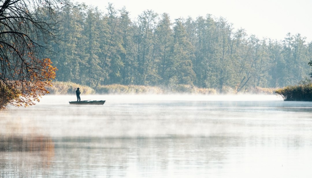 Angler im Boot auf der nebelverhangenen Peene im Herbst, umgeben von herbstlichen B&auml;umen.
