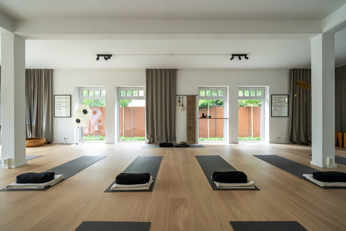 Ein moderner, minimalistischer Yoga- und Meditationsraum mit hellen Holzb&ouml;den, gro&szlig;en Fenstern und dezenter Dekoration. Die Matten sind bereits f&uuml;r eine bevorstehende Yoga-Sitzung vorbereitet, begleitet von weichen Kissen und Handt&uuml;chern. // &copy; Hotel Carpe Diem