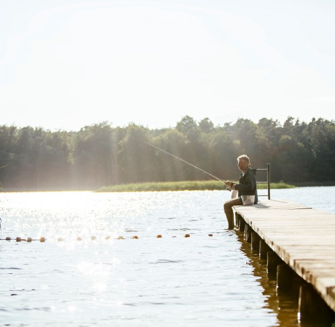Ein Mann sitzt auf einem Holzsteg an einem ruhigen See und angelt, w&auml;hrend die Sonne das Wasser glitzern l&auml;sst.