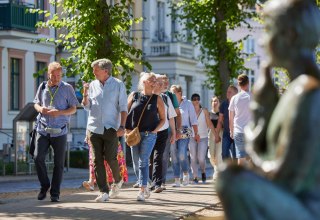 Een groep mensen loopt over een geplaveid pad met een stadsgids. Rechts, onscherp in beeld, is een standbeeld te zien bij de Pfaffenteich-vijver; de villa's verschijnen op de achtergrond. De focus ligt op de stadsgids en een geanimeerd pratend echtpaar. // © Oliver Borchert Een groep mensen loopt over een geplaveid pad met een stadsgids. Rechts, onscherp in beeld, is een standbeeld te zien bij de Pfaffenteich-vijver; de villa's verschijnen op de achtergrond. De focus ligt op de stadsgids en een geanimeerd pratend echtpaar. // © Oliver Borchert