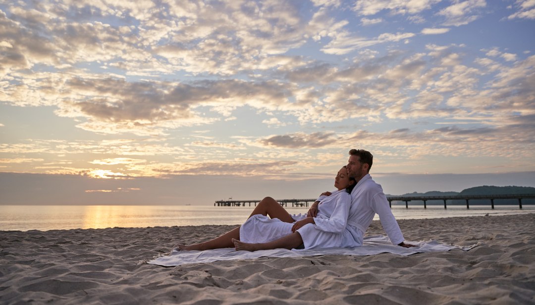 Zwei Personen sitzen am Strand im bademantel mit der Seebrücke Binz im Hintergrund // Ostseefeeling am Strand im Bademantel // © Kurhaus Binz Zwei Personen sitzen am Strand im bademantel mit der Seebrücke Binz im Hintergrund