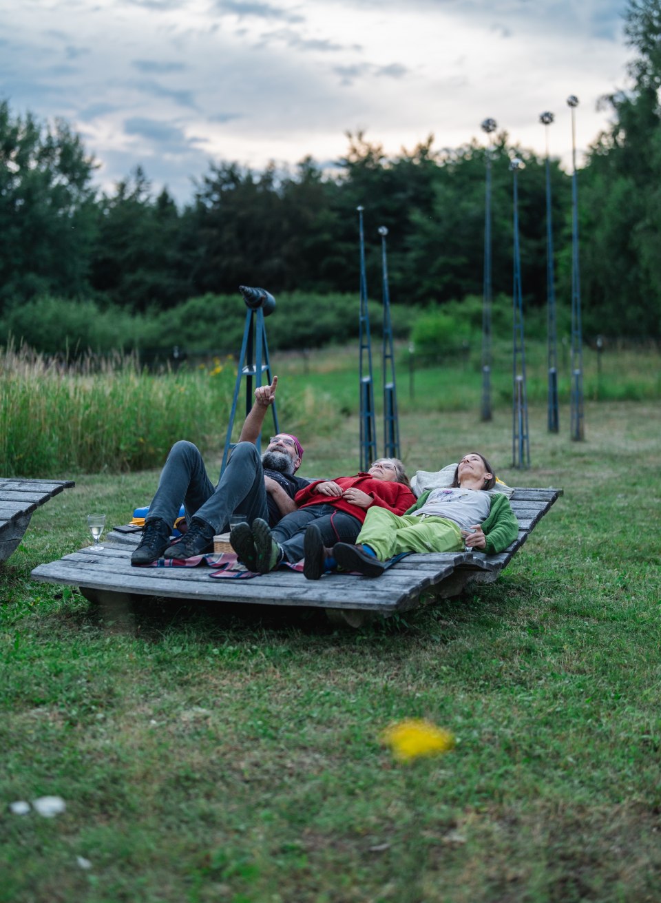 Tre personer ligger p&aring; en tr&aelig;platform midt i det gr&oslash;nne og kigger op mod aftenhimlen i Stjerneparken Mecklenburg-Vorpommern.