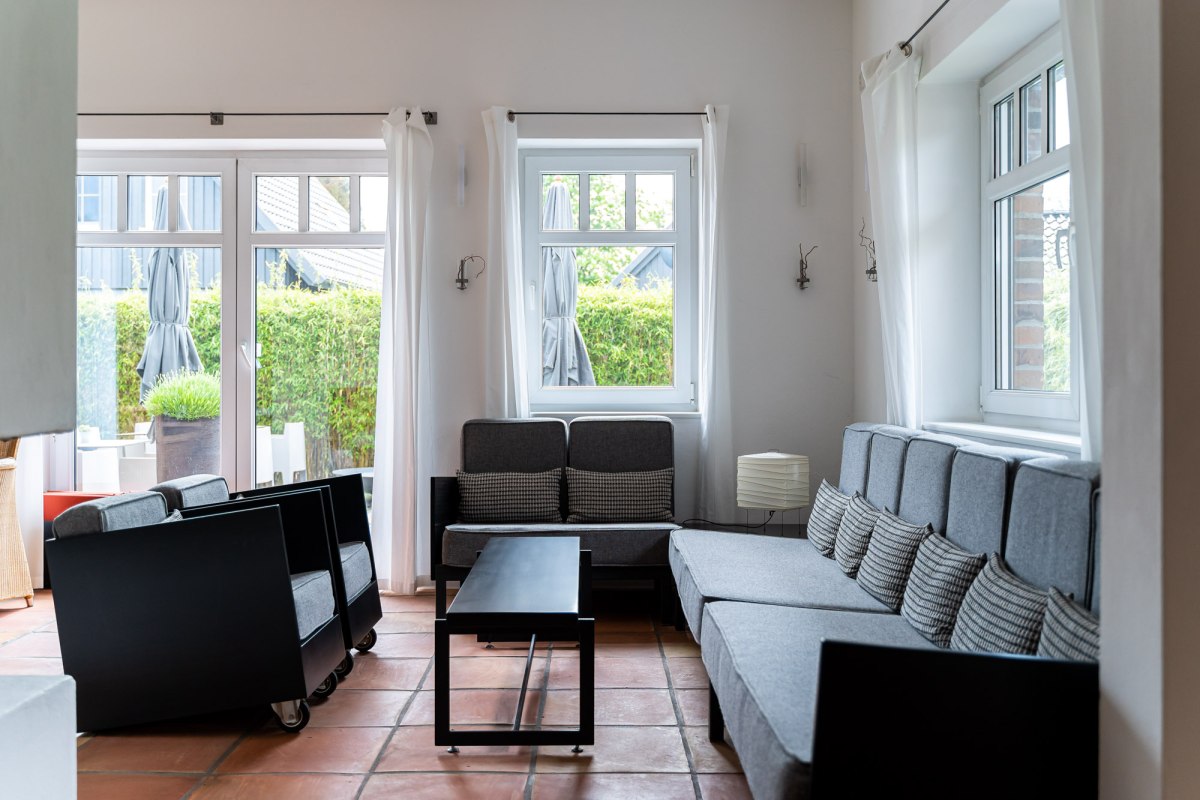 Ein heller und stilvoll eingerichteter Lounge-Bereich mit gro&szlig;en Fenstern, die viel Tageslicht hereinlassen. Graue Sofas und Sessel mit minimalistischen schwarzen Akzenten laden zum Entspannen ein. Die warmen Terrakotta-B&ouml;den und die schlichten wei&szlig;en Vorh&auml;nge verleihen dem Raum eine entspannte und einladende Atmosph&auml;re. // &copy; Hotel Carpe Diem