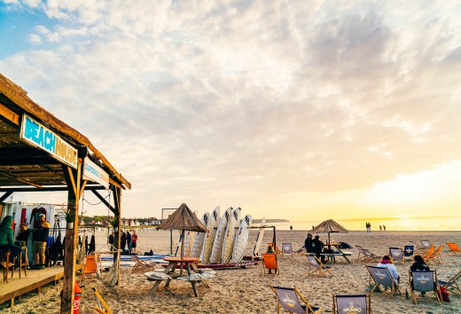 Strandbillede med strandhus, surfbr&aelig;tter og solnedgang i Warnem&uuml;nde, hvor folk slapper af i liggestole.