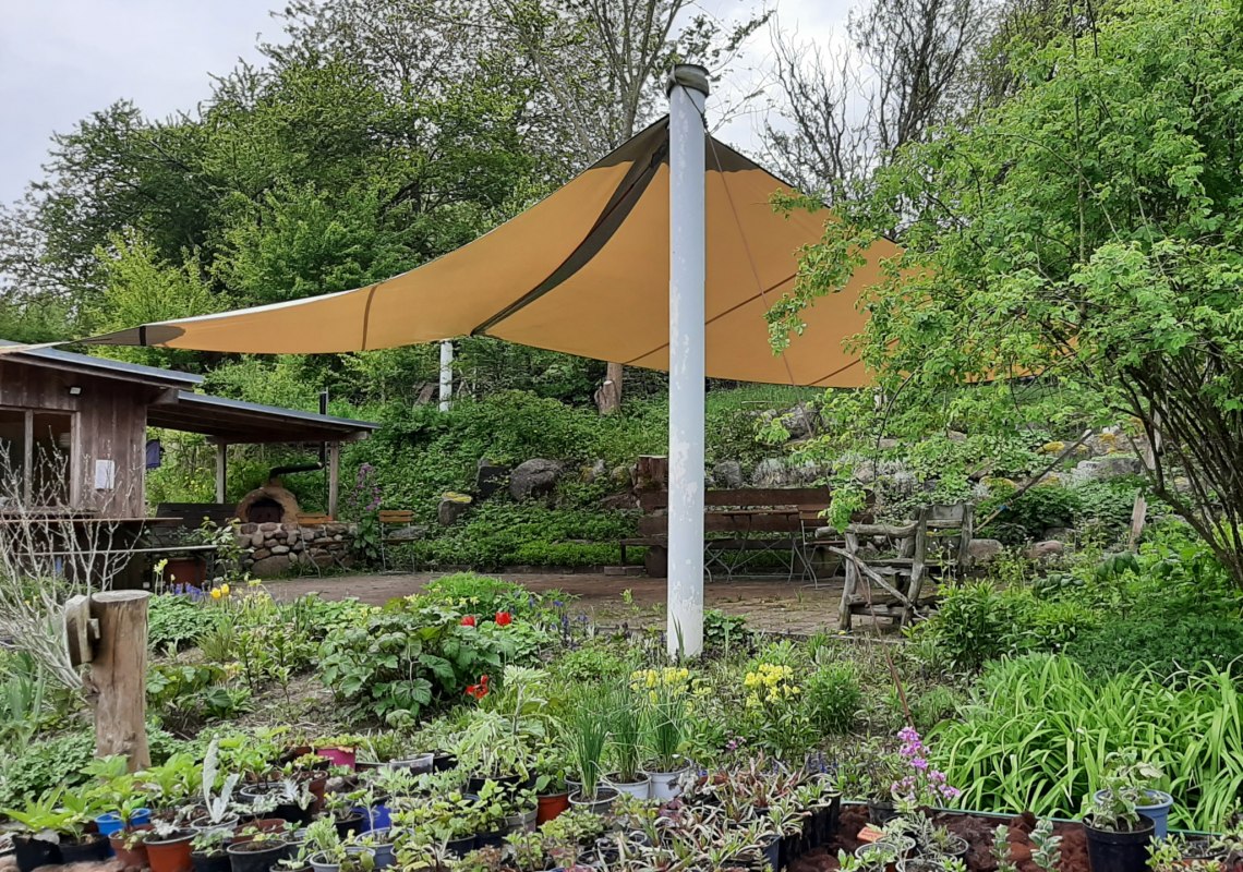 Blick auf die Kiosk-Café-Terasse. Sie ist mit einem großen Segel aus regendichter Plane überdacht. Am unteren Bildrand ist ein Teil vom Jungpflanzentisch zu sehen. // © Mirabell e.V. Blick auf die Kiosk-Café-Terasse. Sie ist mit einem großen Segel aus regendichter Plane überdacht. Am unteren Bildrand ist ein Teil vom Jungpflanzentisch zu sehen. // © Mirabell e.V.