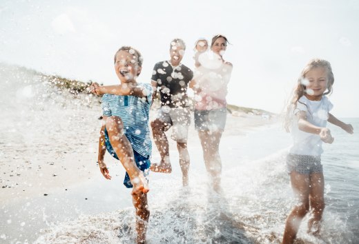 Eine Familie l&auml;uft lachend durch die flachen Ostseewellen am Strand von Dierhagen, w&auml;hrend Wasser in der Sonne lebendig aufspritzt.