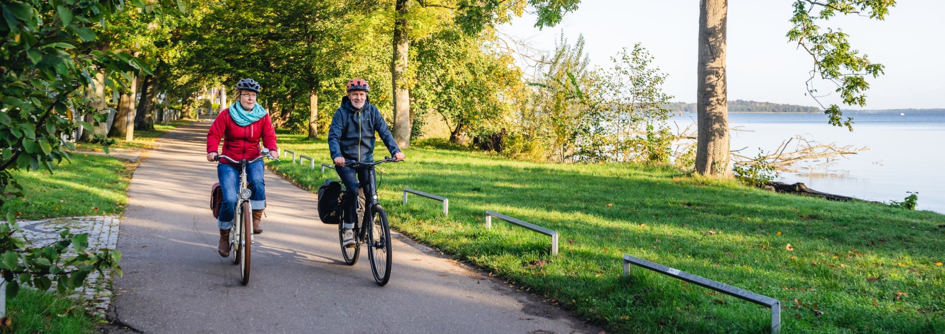 Aktiv am Schweriner See – Fahrradfahren auf dem idyllischen Franzosenweg., © TMV/Gross Zwei Radfahrer auf einem baumgesäumten Weg am Ufer des Schweriner Sees, umgeben von herbstlicher Natur.