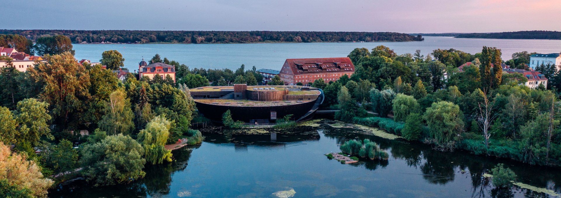 Luftaufnahme des M&uuml;ritzeums in Waren (M&uuml;ritz), umgeben von Wasser und gr&uuml;ner Natur in der Abendd&auml;mmerung.