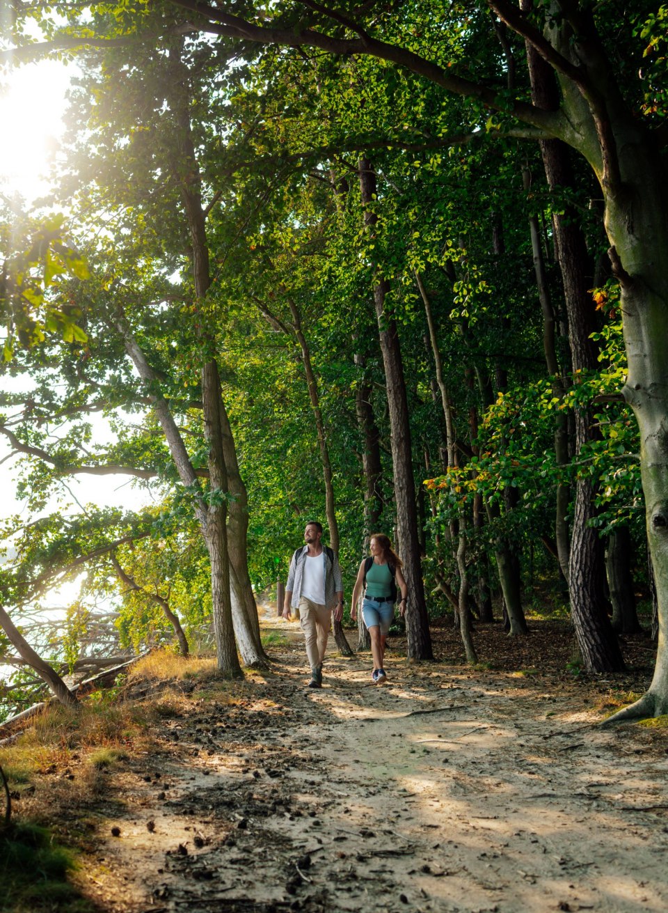 Ein Paar wandert auf einem schattigen Waldweg entlang der K&uuml;ste im Biosph&auml;renreservat S&uuml;dost-R&uuml;gen. Die Sonne scheint durch die B&auml;ume und spiegelt sich im Wasser. Die Atmosph&auml;re ist ruhig und von der nat&uuml;rlichen Sch&ouml;nheit der Umgebung gepr&auml;gt.