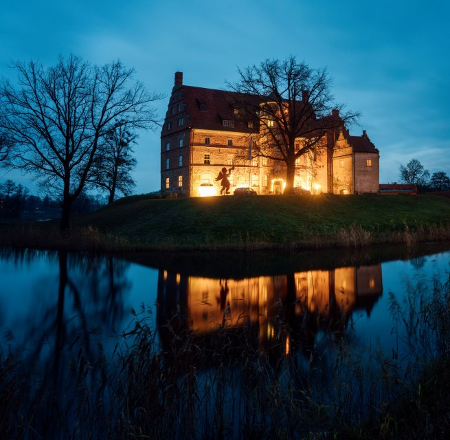 Weihnachtliche Idylle am Schloss Ulrichshusen – festliche Beleuchtung spiegelt sich im Wassergraben und sorgt für eine märchenhafte Abendstimmung., © TMV/Petermann Stimmungsvoll beleuchtetes Schloss Ulrichshusen zur Weihnachtszeit, umgeben von Bäumen und Wassergraben mit Spiegelung im Abendlicht.