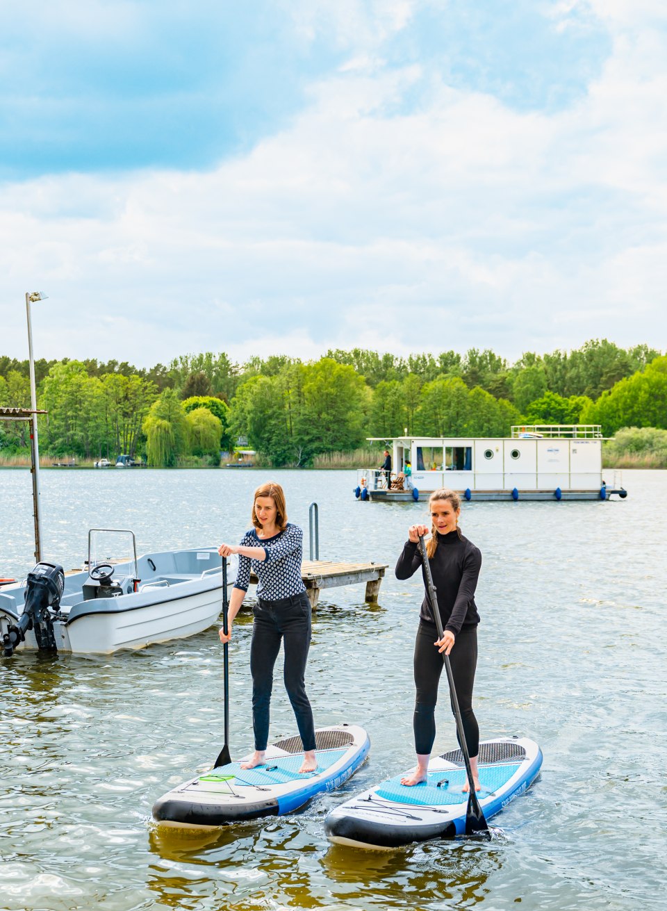 Zwei Frauen paddeln an Land mit SUPs und im HIntergrund ist ein Hausboot // Auf wichtiger Mission: Ausgestattet mit einer wasserdichten Tupperdose paddeln die beiden Freundinnen an Land, um Kuchen zu kaufen. // © MV-T/Tiemann Zwei Frauen paddeln an Land mit SUPs und im HIntergrund ist ein Hausboot