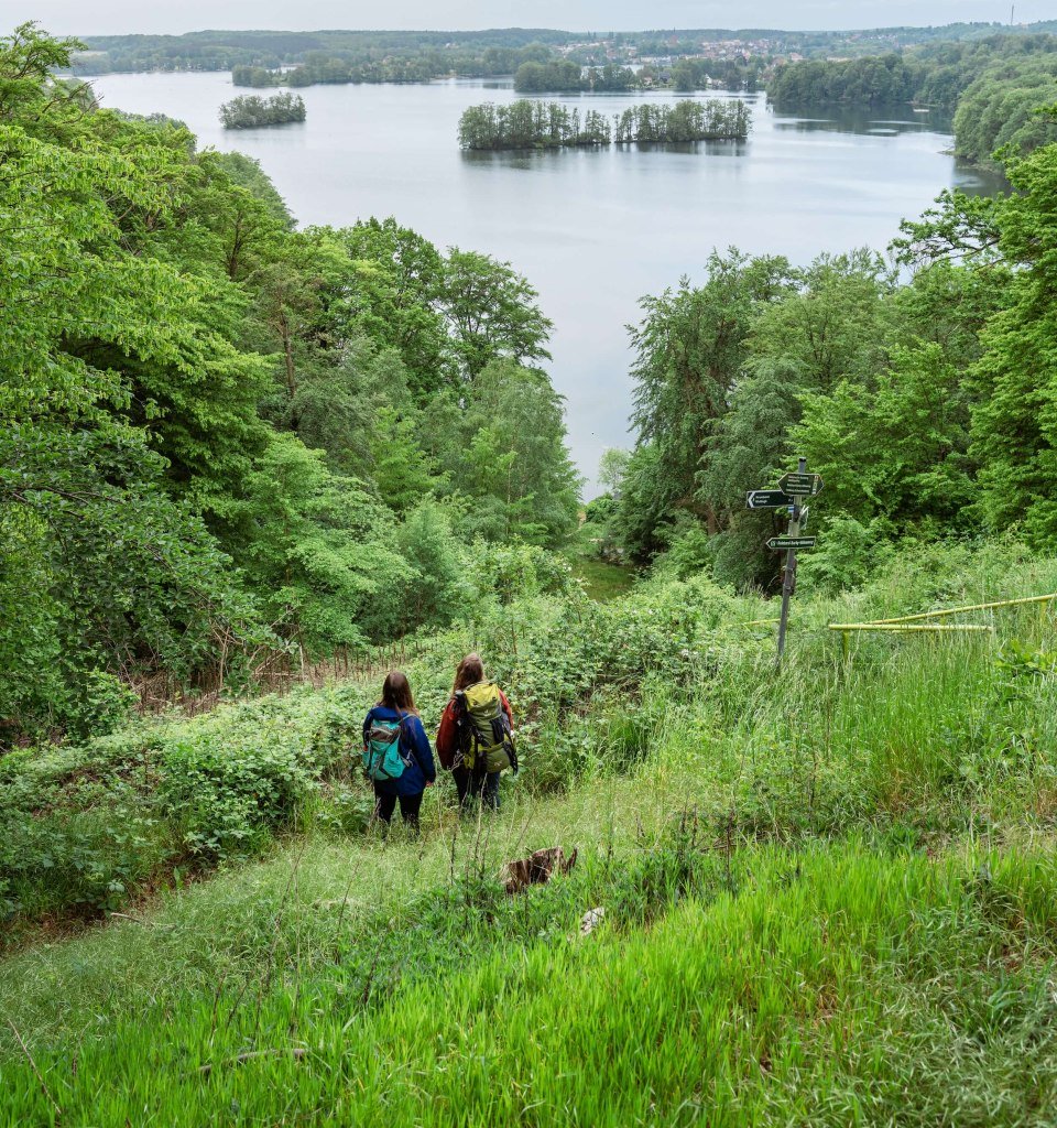 Zwei Frauen wandern auf dem Naturparkweg und schauen vom Berg runter auf den See Schmaler Luzin.