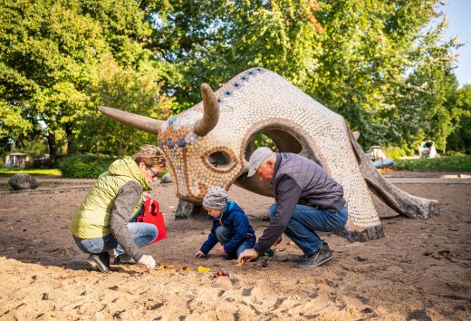 Een vrouw en een man zitten met een kind in het zand voor een betegelde sculptuur van een stier en zoeken kastanjes. // Kastanjes in het zand, bladeren in de hand: op de speeltuin aan het Tollensemeer in Neubrandenburg genieten gezinnen samen van de herfst. // © MV-T/Tiemann Een vrouw en een man zitten met een kind in het zand voor een betegelde sculptuur van een stier en zoeken kastanjes.
