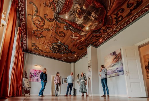 Gäste stehen im Barocksaal und betrachten die reich bemalte Decke mit Ornamenten im Gutshaus Ludorf. // Decke hoch, Blick nach oben: Im Barocksaal im Gutshaus Ludorf erzählen Ornamente von Kunst und Zeit. // © MV-T/Petermann Gäste stehen im Barocksaal und betrachten die reich bemalte Decke mit Ornamenten im Gutshaus Ludorf.