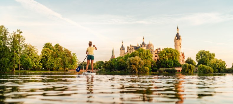 Eine Person paddelt bei warmem Abendlicht auf einem See und blickt auf ein historisches Schloss, umgeben von üppigem Grün. // Beim Gleiten über den Schweriner See entsteht ein stiller, kraftvoller Augenblick, in dem Natur und Schlosskulisse miteinander verschmelzen. // © MV-T/Petermann Eine Person paddelt bei warmem Abendlicht auf einem See und blickt auf ein historisches Schloss, umgeben von üppigem Grün.