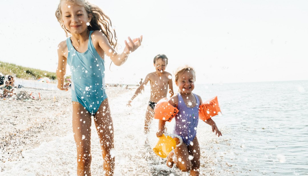 Drei Kinder rennen lachend durch das flache Wasser am Ostseestrand und spritzen fröhlich mit den Wellen. // Unbeschwertes Badevergnügen – lachende Kinder toben am Strand und genießen einen Sommertag an der Ostsee. // © MV-T/Roth Drei Kinder rennen lachend durch das flache Wasser am Ostseestrand und spritzen fröhlich mit den Wellen.