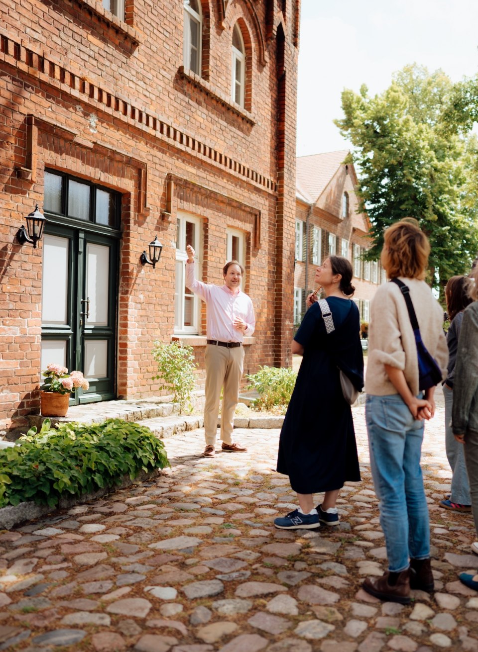 Jannis Achtenhagen erklärt einer Gruppe den Backsteinbau des Gutshauses Ludorf auf einem Kopfsteinpflasterweg im Grünen. // Zwischen Backstein und alten Bäumen führt Jannis Achtenhagen die Gruppe ums Gutshaus Ludorf, zeigt Details an der Fassade und öffnet neue Perspektiven // © MV-T/Petermann Jannis Achtenhagen erklärt einer Gruppe den Backsteinbau des Gutshauses Ludorf auf einem Kopfsteinpflasterweg im Grünen.